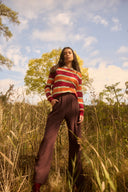 Woman in a colorful striped sweater and brown pants standing in a field with trees and blue sky in the background.