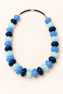 A beaded necklace with alternating large dark navy, light blue, and pale aqua oval beads, finished with a short strand of small dark blue beads and gold-tone clasp, shown on a warm off-white background.