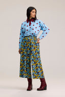 A model stands against a light beige studio background wearing a light blue long-sleeve blouse with red polka dots and a dark red collar, paired with high-waisted wide-leg trousers featuring a blue, yellow, and black floral pattern, and dark red ankle boots.