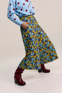 A person wearing a light blue long-sleeve top with red polka dots and a high-waisted, wide-leg skirt with a blue, yellow, and navy floral pattern, paired with burgundy heeled boots, against a light beige studio background.