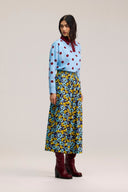 A model stands against a light beige studio background wearing a light blue long-sleeve blouse with red polka dots and a dark red bow collar, paired with a blue floral midi skirt and dark red boots.