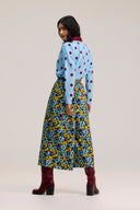 A model is shown from the back wearing a light blue long-sleeve top with red polka dots and a yellow, blue, and navy floral midi skirt, paired with burgundy heeled boots, against a plain light beige studio background.