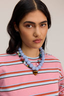 A close-up of a woman wearing a pink, white, and blue striped long-sleeve shirt and a chunky layered beaded necklace with light blue beads and a small gold heart pendant.