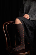 Person wearing brown knee-high socks with orange dots sitting on a wooden chair against a dark background