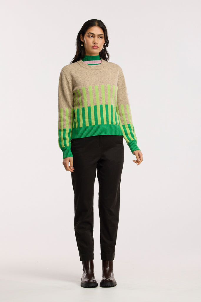 Person wearing a beige sweater with green and light green striped patterns and black pants on a plain background