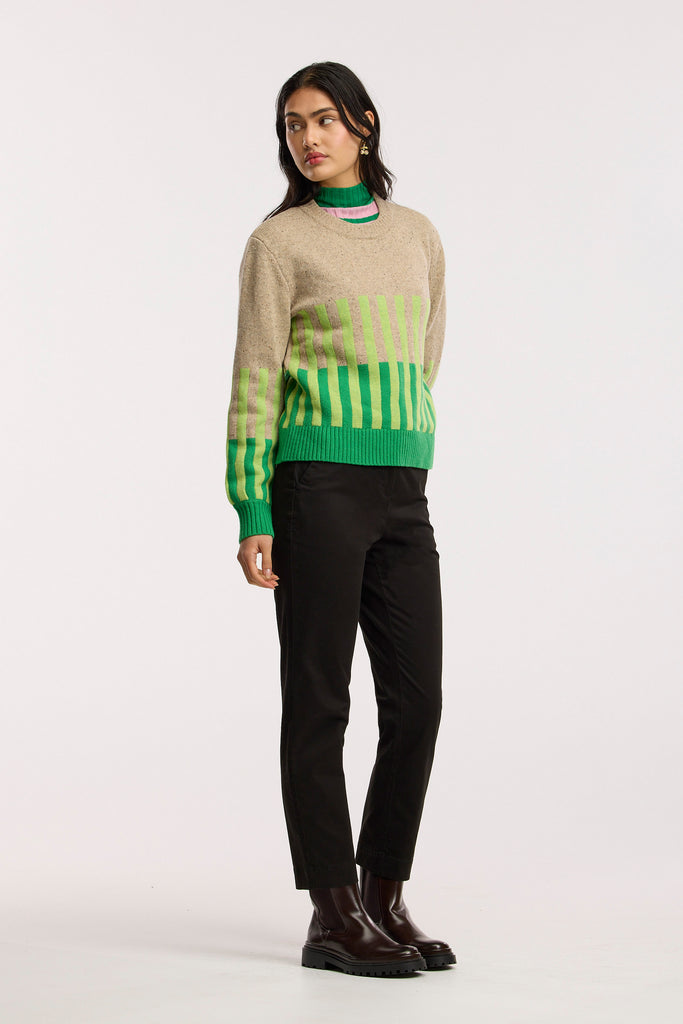 Person wearing a beige sweater with green and light green striped patterns and black pants on a plain background