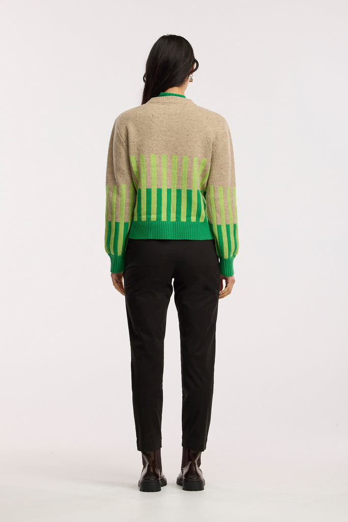 Person wearing a beige sweater with green and light green striped patterns and black pants on a plain background