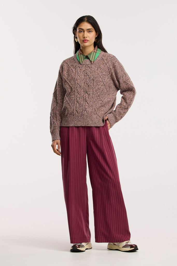 Person wearing a multi-coloured cable knit sweater and red striped pants on a white background