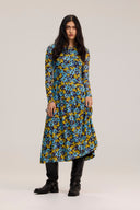 A woman modeling a long-sleeve, knee-length dress with a vibrant blue, yellow, and black floral pattern, paired with black boots, against a plain light gray studio background.