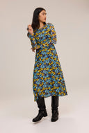A woman modeling a long-sleeved midi dress with a blue, yellow, and black abstract floral pattern, paired with black leather boots, against a plain beige studio background.