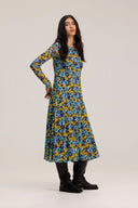 A woman modeling a long-sleeve midi dress with a vibrant blue, yellow, and black floral pattern, paired with black boots, against a plain light beige studio background.