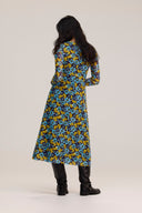 A woman viewed from the back wearing a long-sleeved midi dress with a dense blue, yellow, and black floral pattern, paired with black boots, against a plain light beige studio background.