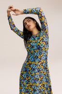 A woman modeling a long-sleeve, high-neck dress with a vibrant blue, yellow, and brown floral pattern against a solid light beige background.