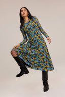 A woman modeling a long-sleeve midi dress with a blue, yellow, and black floral pattern, paired with black knee-high boots, against a solid light beige background.