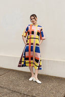 A woman stands outdoors against a light beige wall wearing a short-sleeve midi dress with a bold multicolor geometric stripe pattern in pink, yellow, blue, orange, black, and brown. She is wearing white pointed flats.
