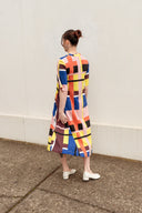 A woman stands with her back to the camera wearing a short-sleeved midi dress with a colorful geometric block pattern in yellow, orange, blue, pink, black, and white. She is standing on a concrete sidewalk against a plain off-white wall.