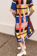 A person wearing a short-sleeve midi dress with a bold geometric plaid pattern in blue, pink, yellow, brown, black, and white, paired with white low-heeled shoes, standing on a concrete surface against a white wall.