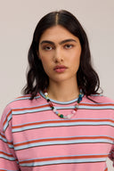 A woman with dark hair wearing a pink crewneck sweatshirt with horizontal brown, white, and black stripes, accessorized with a colorful beaded necklace, set against a plain light beige background.