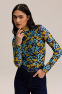 A woman modeling a long-sleeve mock-neck shirt with a vibrant blue and yellow floral pattern on a dark background, paired with dark blue jeans and a dark belt, against a solid light beige background.