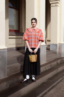 A woman stands on stone steps in front of a building, wearing a bright orange and red plaid puff-sleeve top, a long black skirt, white socks, and dark loafers, while holding a small woven handbag.