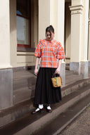 A woman stands on the steps of a beige building wearing a bright orange and white plaid short-sleeve blouse with a large collar, a long black skirt, white socks, and dark loafers. She is holding a small woven straw handbag.