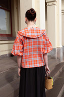 A woman viewed from behind wearing a bright orange and white plaid blouse with a large ruffled collar and puff sleeves, paired with a long black skirt. She is standing on stone steps in front of a cream-colored building, holding a small woven straw handbag.