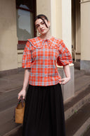 A woman stands on outdoor stone steps wearing a short-sleeve orange and light blue plaid blouse with a large ruffled collar and puff sleeves, paired with a long black skirt. She holds a woven straw handbag.
