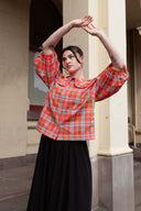 A woman posing outdoors in front of a beige stone building, wearing a red and blue plaid long-sleeve shirt with a Peter Pan collar and puff sleeves, paired with a long black skirt.