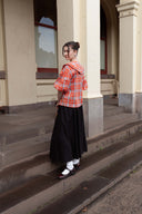 A woman stands on outdoor stone steps in front of a beige building, wearing a red and orange plaid hooded jacket, a long black skirt, white socks, and black-and-white Mary Jane shoes.