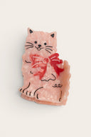 A pink cat-shaped hair claw clip with a red bow detail and black line art accents, shown on a plain off-white background.