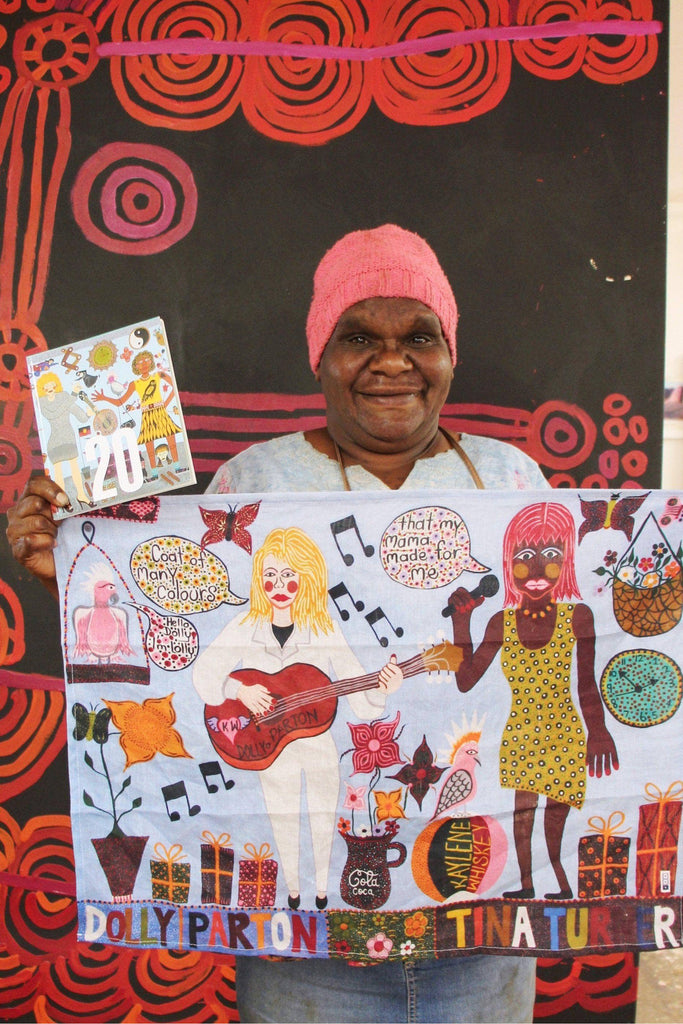 Photo of artist Kaylene Whiskey holding a colorful tea towel featuring illustrations of Dolly Parton and Tina Turner